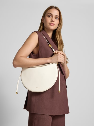 Frauenmodel trägt Handtasche aus Leder mit Label-Detail von FURLA in Hellbeige, goldfarbene Details, Schulterriemen, Ganzkörperansicht von vorne. Hauptfach mit Magnetverschluss, Breite 29 cm, Länge 26 cm, Tiefe 3 cm, Länge Schulterriemen 40 cm.