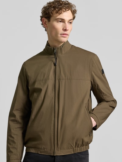 Männermodel trägt Jacke mit Stehkragen Modell 'Calvin' von Brax in Olivgrün, Baumwoll-Mix mit Stretch, Reißverschlusstaschen, Ganzkörperansicht von vorne.