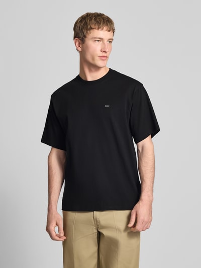 Männermodel trägt T-Shirt mit Label-Detail und Rundhalsausschnitt von Dickies, reine Baumwolle, Relaxed Fit, Ganzkörperansicht von vorne.