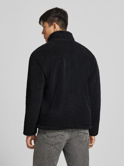 Männermodel trägt Fleecejacke mit Label-Detail und Reißverschluss von REVIEW in Schwarz, Teddyfell-Struktur, Ganzkörperansicht Rückansicht. Regular Fit, Stehkragen, Rückenlänge bei Größe M: 70 cm, Schulterbreite: 56 cm, Brustweite: 59 cm, Ärmellänge: 64 cm.