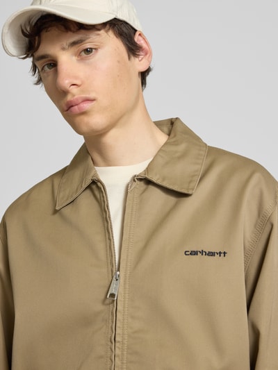 Mężczyznamodel nosi Jacke mit Label-Stitching und Eingrifftaschen od Carhartt Work In Progress w kolorze beżowym, krój regular fit, wykładany kołnierz, zamek błyskawiczny, wpuszczane kieszenie, wyhaftowane logo, szerokość ramion w rozmiarze M: 59 cm, szerokość w rozmiarze M: 60 cm, długość tyłu w rozmiarze M: 68 cm, długość rękawów w rozmiarze M: 20 cm, detal struktury gładkiej tkaniny, zbliżenie na górną część ciała, przód