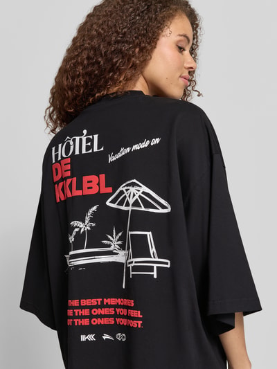 Frauenmodel trägt Cropped T-Shirt mit Statement-Print Modell 'Vacation' von Karo Kauer, Reine Baumwolle, Oversized-Fit, Rückenlänge 62 cm, schwarze Grundfarbe mit weiß-rotem Print, Ganzkörperansicht Rückansicht.