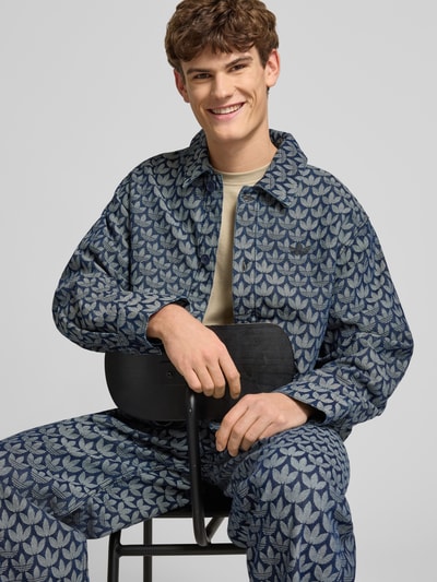 Męski model nosi Regular Fit Jacke in Denim-Optik Modell 'Monogram' od adidas Originals, bawełna, krój regular fit, niebieska, wzór monogram, z długimi rękawami i kołnierzem kent, guziki, mankiety sportowe, długość tyłu 72 cm, szerokość ramion 50 cm, szerokość w rozmiarze M 59 cm, długość rękawa 66 cm, na tle jasnym, zbliżenie na górną część ciała, widok z przodu.