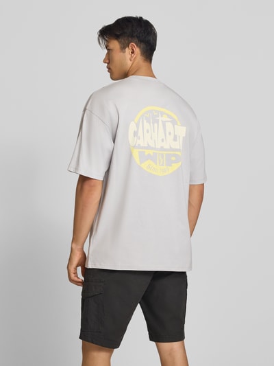Männermodel trägt Relaxed Fit T-Shirt aus reiner Baumwolle Modell 'Luggage' von Carhartt Work In Progress in Beige mit großem gelbem Rückenprint, lockere Passform, schwarze Shorts, ganzkörperansicht Rückansicht, Herren T-Shirt Relaxed Fit aus reiner Baumwolle, Label-Detail, Rückenlänge Größe M 78 cm.