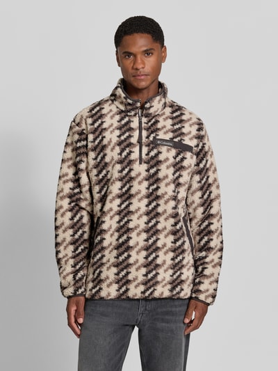 Męski model nosi Regular Fit Pullover mit Logo-Patch od Columbia, Polar, krój regular fit, naszywka z logo, kołnierz zapinany, Długość tyłu w rozmiarze M: 70 cm, Długość rękawów w rozmiarze M: 68 cm, beżowo-czarne wzory, tekstura puszysta, widok całej sylwetki z przodu.