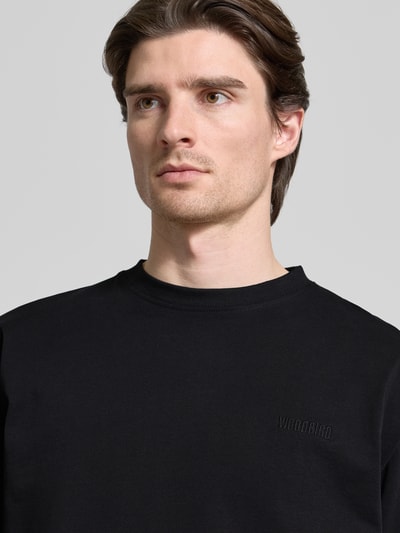 Męski model nosi Longsleeve mit Label-Stitching Modell 'Baine' von WOODBIRD, czarna bawełna organiczna, krój regular fit, dzianina, okrągły dekolt, wykończenia prążkowane, długość tyłu w rozmiarze M: 71 cm, zbliżenie na górną część ciała.