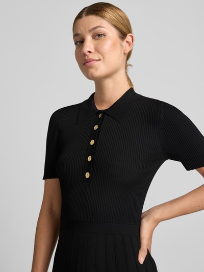 Frauenmodel trägt Polokleid aus Viskose-Mix mit Knopfleiste von MICHAEL Michael Kors in Schwarz, Ripp-Optik, Nahaufnahme Oberkörper. Midikleid mit Stretch-Anteil, A-Linie, goldfarbene Knöpfe, Kontext: Rückenlänge 114 cm, Brustweite 40 cm, Ärmellänge 24 cm bei Größe S.