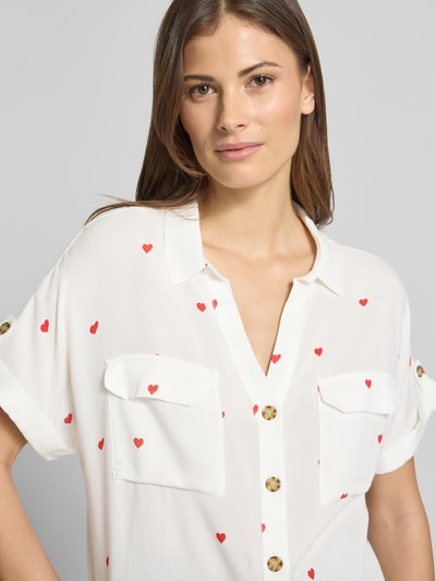 Kobiece modelka nosi Comfort Fit Bluse aus reiner Viskose Modell 'BUMPY' von Vero Moda, biała bluzka z czerwonym wzorem serc, kieszenie na piersi, krótki rękaw z mankietem, lekka tkanina, krój comfort fit, kołnierz koszulowy, zapięcie na guziki, materiał 100% wiskoza, widok na górną część ciała z przodu.