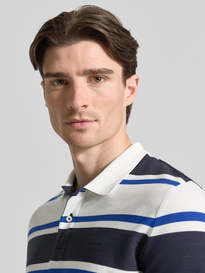 Männermodel trägt Slim Fit Poloshirt mit Logo-Stitching von Gant, weiß-blaue Streifen, reine Bio-Baumwolle, Nahaufnahme Oberkörper.