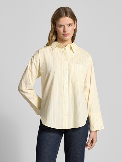 Vrouwenmodel draagt Relaxed Fit Hemdbluse met borstzak Model 'Poplin' van Gina Tricot in geel met witte strepen, katoenen stof, volledige voorkant, lange mouwen, sportmanchetten, afgeronde zoom, borstzak, overhemdkraag, context: relaxed fit, pure katoen, lengte 72 cm, mouwlengte 56 cm, vooraanzicht bovenlichaam.