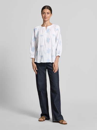 Frauenmodel trägt Bluse aus Viskose-Mix mit verlängertem Rücken von Apricot, weiß-blau, Rundhalsausschnitt, Loose-Fit, Ganzkörperansicht von vorne. Damen Bluse aus Viskose-Baumwoll-Mix, kurze Knopfleiste, Rückenlänge 68 cm, Ärmellänge 52 cm, Größe S.