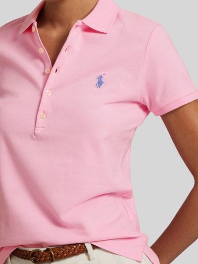 Kobiece modelka ma na sobie Slim Fit Poloshirt aus Baumwoll-Mix od Polo Ralph Lauren, jednolity róż, wyhaftowane logo, kołnierzyk polo, listwa guzikowa, krótki rękaw, materiał pika, mieszanka bawełny, z rozcięciem u dołu, krój slim fit, widok z przodu, zbliżenie na górę ciała.