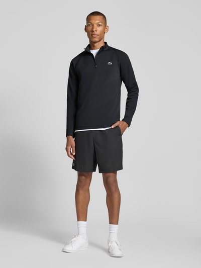 Męskki model nosi Regular Fit Sport-Langarmshirt mit Ultra-Dry-Technologie od Lacoste w kolorach czarnym i białym, sportowa faktura tkaniny, Ganzkörperansicht von vorne.