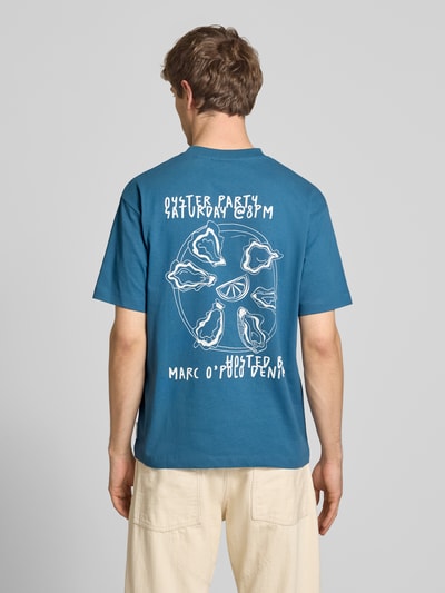 Männermodel trägt Relaxed Fit T-Shirt aus reiner Baumwolle mit Motiv-Print von Marc O'Polo Denim, Optik: Unifarben, Passform: Relaxed-Fit, Motiv-Print, Rundhals, Ärmelform: Kurzarm, Material: reine Baumwolle, kräftiges Blau mit weißem Print. Ganzkörperansicht Rückansicht.