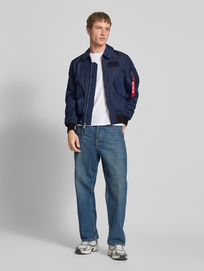 Männermodel trägt Regular Fit Bomberjacke mit Logo-Patch von Alpha Industries in Dunkelblau, Umlegekragen, AufgesetzteTaschen, Reißverschluss, elastische Abschlüsse, Rückenlänge bei Größe M: 66 cm, Schulterbreite 44 cm, Brustweite 56 cm, Ärmellänge 66 cm, Ganzkörperansicht von vorne.
