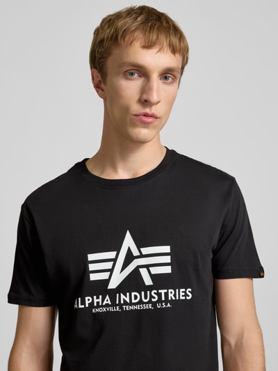 Männermodel trägt T-Shirt mit Logo und Rundhalsausschnitt von Alpha Industries, schwarze Baumwolle, Regular Fit, Nahaufnahme Oberkörper.