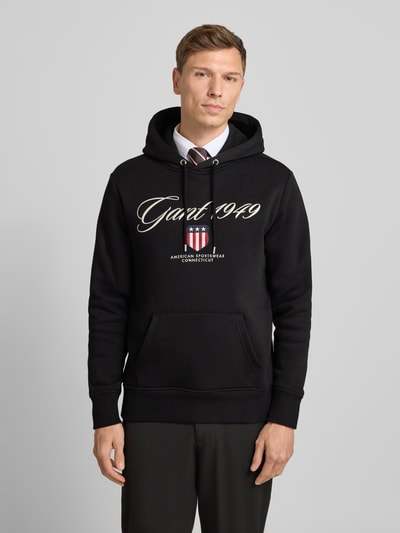 Männermodel trägt Hoodie mit Label-Stitching und Känguru-Tasche von Gant, Baumwoll-Mix, Regular-Fit, schwarze Farbe, Nahaufnahme Oberkörper.