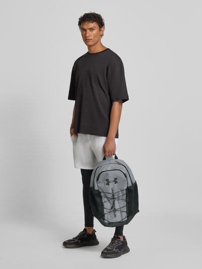 Mannelijk model draagt Rucksack mit Tragehenkel van Under Armour in grijs met labelprint, hoofvak met ritssluiting, draagband, verstelbare schouderbanden, zijvak met ritssluiting, breedte 31 cm, lengte 48 cm, diepte 18 cm, bandlengte 53 cm, ganzkörperansicht van voren.