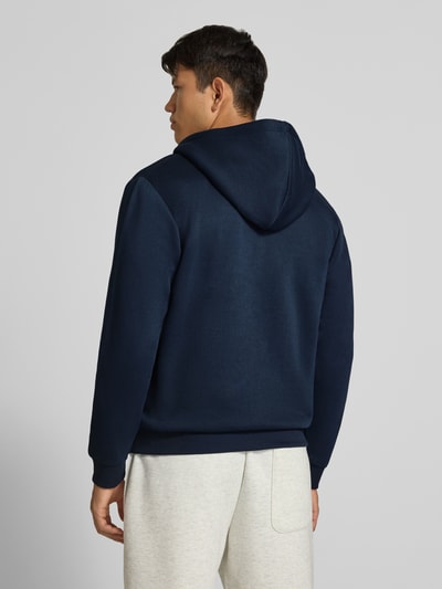 Männermodel trägt Regular Fit Sweatjacke aus Baumwoll-Mix Modell 'BRADLEY' von JACK & JONES in Dunkelblau, Ganzkörperansicht Rückansicht. Kapuze mit Tunnelzug, elastische Abschlüsse, geteilter Känguru-Tasche, Herren Sweatjacke, Rückenlänge 67 cm, Ärmellänge 68 cm.