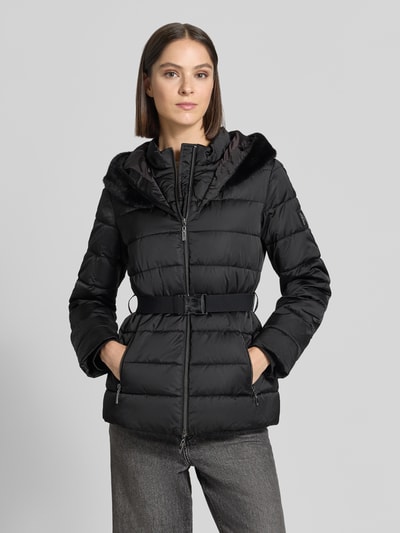 Modelka nosi 'Jacke in Stepp-Optik' od monari, czarny kolor, taliowany krój slim fit, kaptur z ekofutrem, wypełniony kołnierz, dwukierunkowy zamek, długość tyłu 68 cm, szerokość ramion 46 cm, szerokość w rozmiarze 36: 50 cm, długość rękawa 63 cm, całopostaciowy widok z przodu.