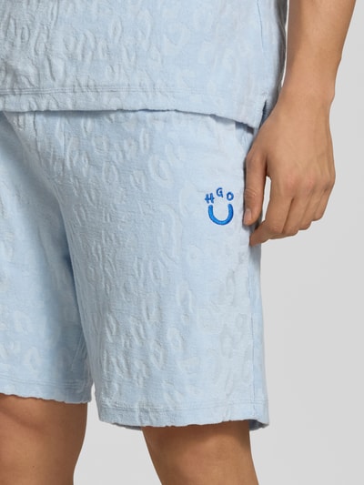 Mężczyznamodel nosi Regular Fit Shorts aus Frottee od HUGO w jasnoniebieskim kolorze, logo stitching, tekstura frotte, boczne kieszenie, tylna naszywana kieszeń, krój regular fit, zbliżenie na dolną część ciała.