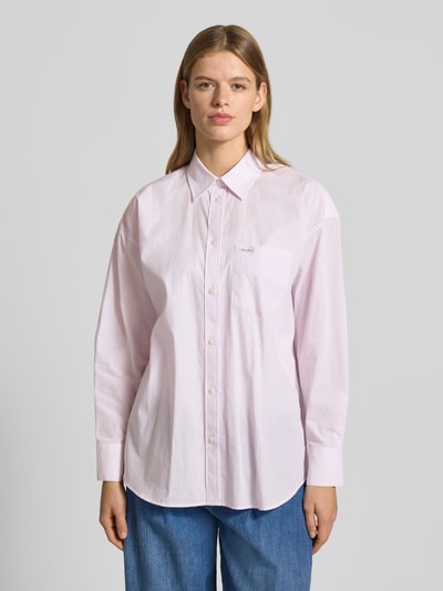Frauenmodel trägt Oversized Hemdbluse mit Brusttasche von Guess Jeans in zartem Rosa, reine Baumwolle, Logo-Patch, Knopfleiste, Ganzkörperansicht von vorne.