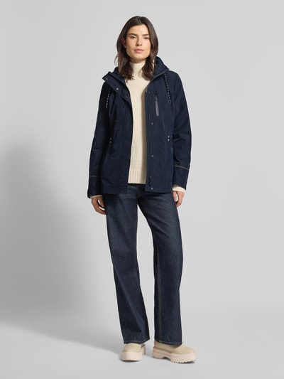 Frauenmodel trägt dunkelblaue Funktionsjacke mit Brusttasche von Gil Bret, taillierte Passform, Kapuze, Stehkragen, Tunnelzug, Reißverschluss, Rückenlänge 74 cm, Schulterbreite 41 cm, Brustweite 47 cm, Ärmellänge 65 cm, Ganzkörperansicht von vorne.