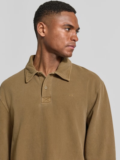 Męski model nosi Loose Fit Poloshirt aus Baumwolle Modell 'Thiago' od Wood Wood w odcieniu khaki, faktura bawełniana, logo na piersi, krój loose fit, polski kołnierzyk, czysta bawełna; kontekst: długość tyłu w rozmiarze M: 71 cm, zbliżenie na górną część tułowia