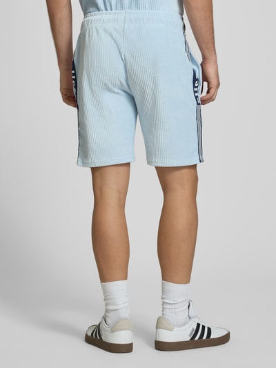 Męski model nosi jasnoniebieskie szorty z prążkowanego sztruksu, elastyczny pas, model 'TOMATRO' od Ellesse, krój regular fit, szerokość w rozmiarze M 42 cm, długość nogawki wewnętrznej 21 cm, widok całej sylwetki z tyłu