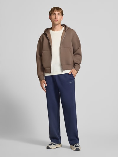 Männermodel trägt Relaxed Fit Sweatjacke mit Zweiwege-Reißverschluss von JACK & JONES, braun, Känguru-Tasche, Ganzkörperansicht von vorne. Elastischer Material-Mix, elastische Abschlüsse, Rückenlänge 65 cm, Ärmellänge 65 cm, lässig kombiniert, glatte Textur.