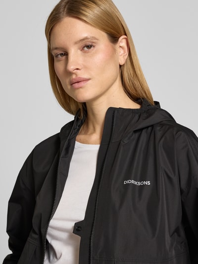 Vrouwenmodel draagt Jacke mit Pattentaschen Modell 'MILENA' van Didriksons in zwart met labelprint, recht pasvorm, capuchon, mouwsluitingen, ritssluiting, tunnelkoord in band, detailopname bovenlichaam.