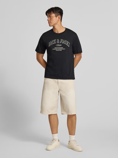 Männermodel trägt Regular Fit T-Shirt aus reiner Baumwolle Modell 'Denver' von JACK & JONES, schwarzes Shirt, heller Print, beige Shorts, Ganzkörperansicht von vorne. Herren T-Shirt mit Logo-Print, Rückenlänge bei Größe M: 69 cm.