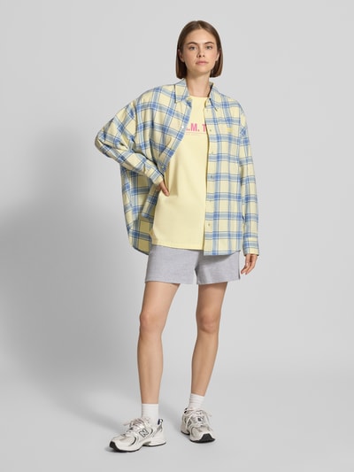 Frauenmodel trägt T-Shirt mit Rundhalsausschnitt Modell 'AM to PM' von OH APRIL in Gelb mit Print aus reiner Baumwolle, Oversized, darunter kurze graue Hose, darüber kariertes Hemd in Blau-Gelb, Ganzkörperansicht von vorne.