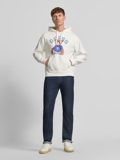 Männermodel trägt Hoodie mit Label-Print und Känguru-Tasche von GUESS in Weiß, Baumwoll-Mix mit Stretch-Anteil, Regular-Fit, Ganzkörperansicht von vorne.
