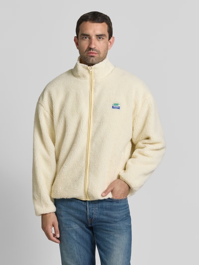 Männermodel trägt fleecejack met opstaande kraag en labeldetail model 'HOKTOWN' van American Vintage in offwhite, relaxed fit, ritssluiting, steekzakken, context: heren fleecejack, ruglengte bij maat M/L 69 cm, schouderbreedte 56 cm, borstwijdte 48 cm, mouwlengte 55 cm, ganzkörperansicht van voren.