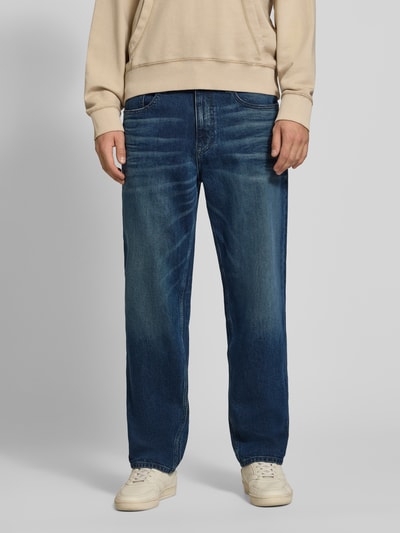 Männermodel trägt Tapered Fit Jeans aus Baumwoll-Mix von Tommy Jeans in Mittelblau mit leichter Waschung, Ganzkörperansicht von vorne. Herren Tapered Fit Jeans von Tommy Jeans.