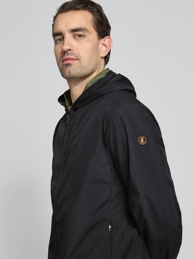 Männermodel trägt schwarze Jacke mit Zweiwegereißverschluss Modell 'Zayn' von SAVE THE DUCK, glatte Textur, Nahaufnahme Oberkörper. Regular Fit, Eingrifftaschen mit Reißverschluss, Kapuze, Details aus dem Kontext sichtbar.