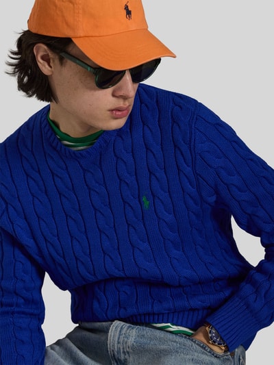 Męski model nosi Regular Fit Strickpullover aus reiner Baumwolle od Polo Ralph Lauren, jednokolorowy niebieski, ścieg warkoczowy, imitacja dzianiny, detal z logo, okrągły dekolt, bawełna, długi rękaw, prążkowane wykończenia, Ganzkörperansicht z przodu.