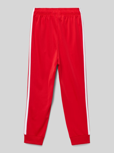 Produktfoto, Trainingshose mit Label-Stitching von adidas Originals für kinder, rot mit weißen Streifen, Regular Fit, elastischer Bund, Eingrifftaschen, Innenbeinlänge 34 cm bei Größe 140, glatte Stoffstruktur.