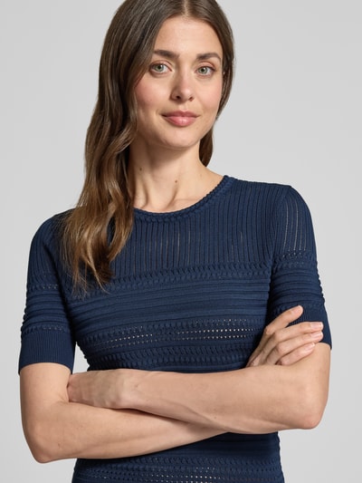 Frauenmodel trägt T-Shirt in Strick-Optik mit Rundhalsausschnitt von Guess in Dunkelblau mit gerippten Abschlüssen, Nahaufnahme Oberkörper. Körpernaher Schnitt, Material-Mix mit Viskose-Anteil, Kurzarm.