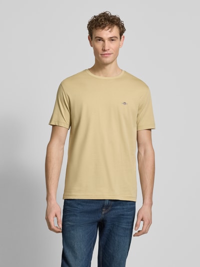 Męski model nosi Regular Fit T-Shirt mit Logo-Stitching von Gant z bawełny, beżowy kolor, okrągły dekolt, długość tyłu 68 cm w rozmiarze M, Ganzkörperansicht von vorne.