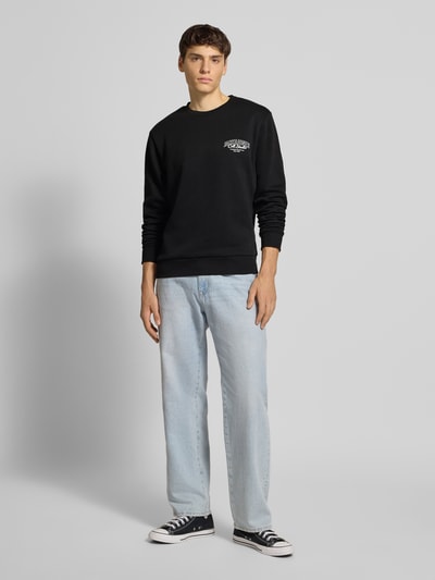 Męski model nosi bluzę z nadrukiem z logo model 'MAKOTO' od Jack & Jones, mieszanka bawełny, krój regular fit, okrągły dekolt, prążkowane wykończenia, czarna, gładka faktura, długość tyłu 62 cm, długość rękawów 70 cm, widok całej sylwetki z przodu.