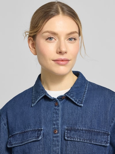 Vrouwenmodel draagt Cropped Jeansbluse aus reiner Baumwolle Modell 'POSEY' van Vero Moda in denimoptiek met hemdblousekraag, boxy fit, zichtbare borstklepzakken, lange mouw met manchet, eenrijige knoopsluiting, textuur zuiver katoen, detailopname bovenlichaam.