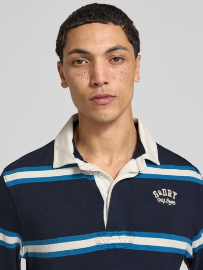 Männermodel trägt Poloshirt aus reiner Baumwolle von Superdry in Dunkelblau mit weißen und blauen Streifen, Nahaufnahme Oberkörper. Gerader Schnitt, Polokragen mit Abdeckleiste, Label-Stitching, elastische Abschlüsse, Rückenlänge bei Größe M 71 cm.