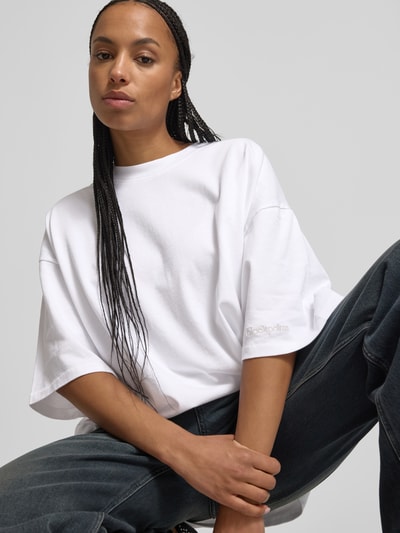Frauenmodel trägt Oversized T-Shirt mit Label-Stitching von black palms, reine Baumwolle, Rundhalsausschnitt, Halbarm, weiß, Nahaufnahme Oberkörper.