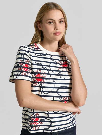 Frauenmodel trägt Regular Fit T-Shirt Modell 'FLOWER GRAPHIC REG C-NK S' von Tommy Hilfiger mit Optik:Allover-Muster, Rundhals, Kurzarm, reine Baumwolle, Farben Weiß, Dunkelblau und Rot, Grafik-Muster und Blumen-Muster, Nahaufnahme Oberkörper.