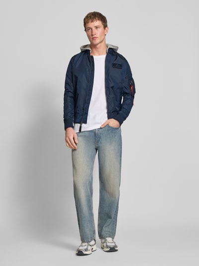 Männermodel trägt Herren Blouson mit Logo-Anhänger Modell 'MA-1 TT' von Alpha Industries, dunkelblau mit Kapuze, elastische Abschlüsse, Label-Patch, Armtasche, Ganzkörperansicht von vorne. Rückenlänge 69 cm, Schulterbreite 46 cm, Brustweite 54 cm, Ärmellänge 67 cm.