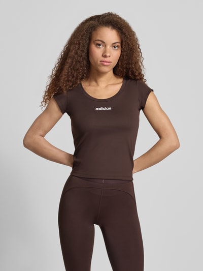 Vrouwenmodel draagt Slim Fit Sport T-Shirt met Label Print van adidas Sportswear in effen bruin, stretch geweven materiaal, korte mouwen, ronden hals, nahaufnahme bovenlichaam.