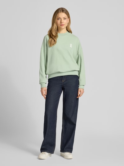 Modelka kobieca ma na sobie Oversized Fit Sweatshirt aus reiner Bio-Baumwolle Modell 'ICONIC A ALIZAA' od Armedangels, gładki jasny zielony, długi rękaw, logo na piersi, okrągły dekolt, bawełna organiczna, tekstura miękka, widok całej sylwetki z przodu.