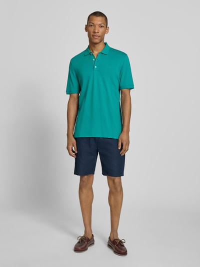 Męski model nosi Slim Fit Poloshirt aus Baumwoll-Mix Modell 'Pete' von Brax, elastyczna mieszanka bawełny, krój slim fit, kołnierzyk polo, długość tyłu w rozmiarze M: 71 cm, kolor turkusowy z granatowymi szortami, pełny widok z przodu.